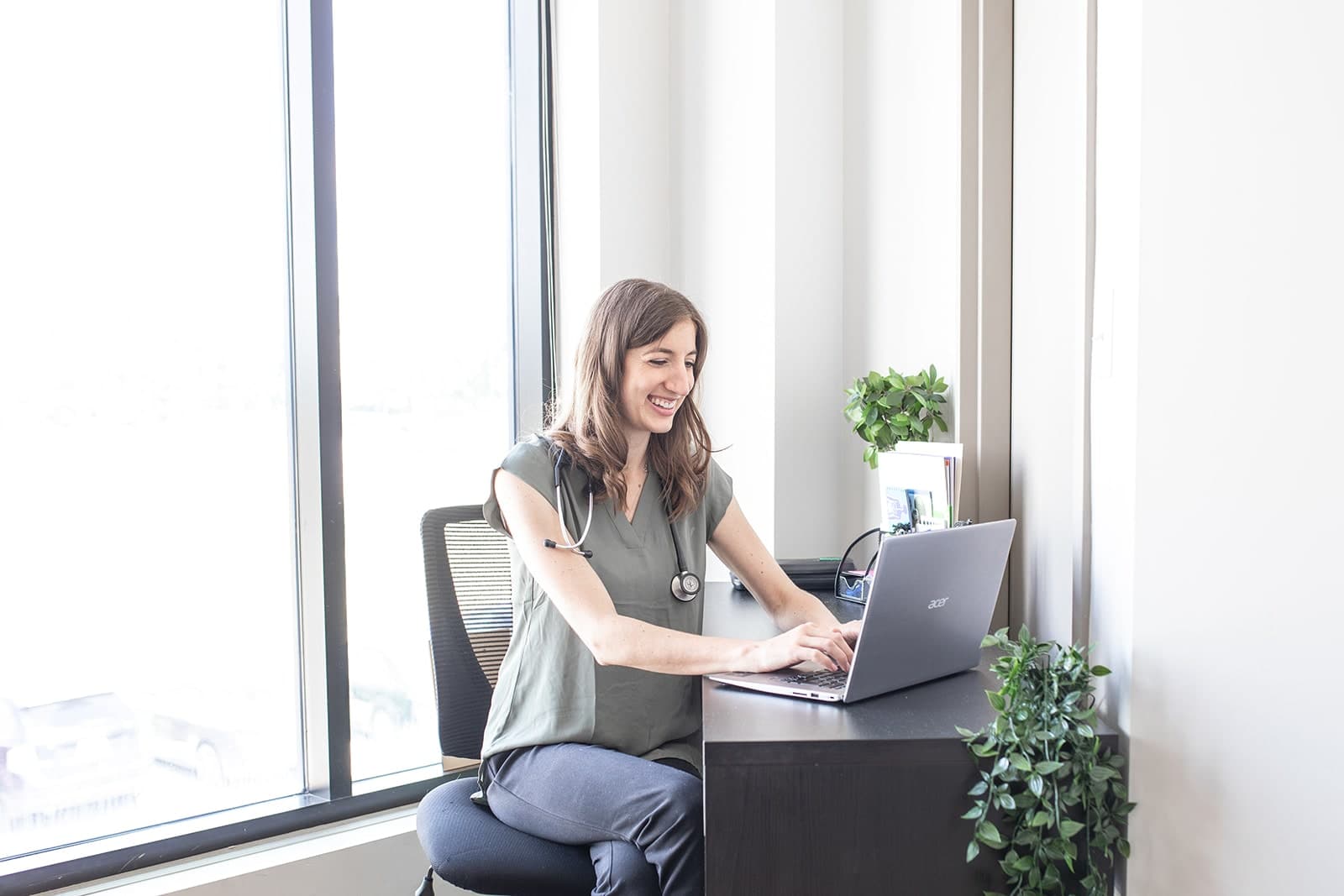 Healthcare practitioner in grey scrubs with stethoscope, working at laptop in bright office with floor-to-ceiling windows and potted plants
