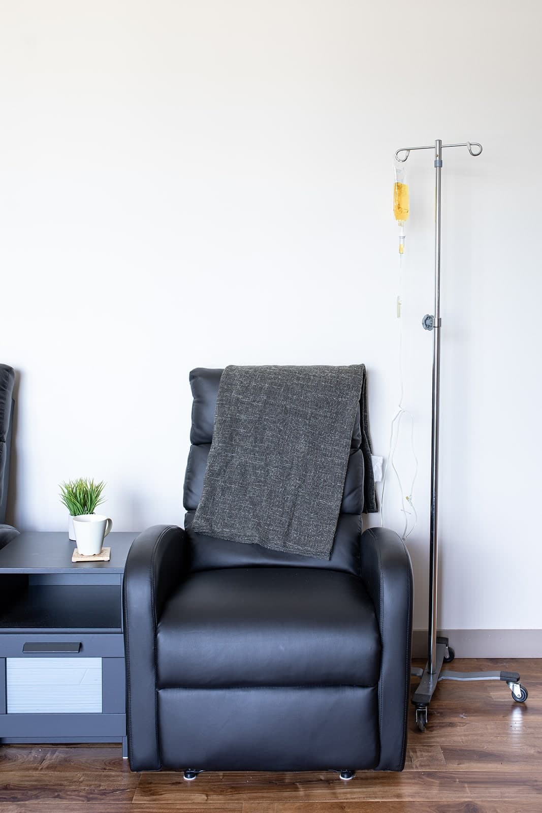 IV therapy treatment chair with gray blanket, IV stand with yellow bag, small side table with potted plant, hardwood floor, white wall, Burlington