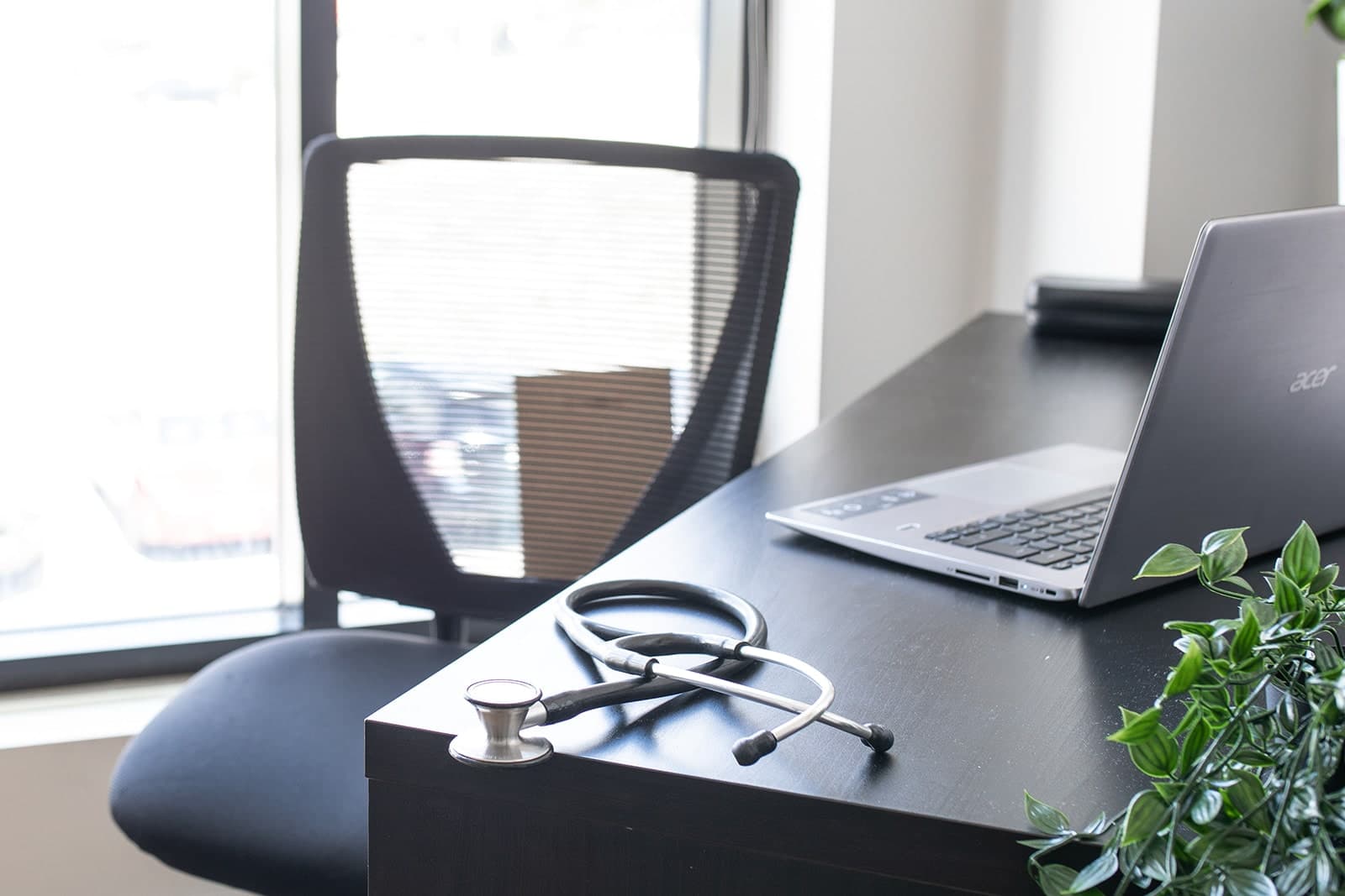 Medical office desk with stethoscope, laptop, and potted plant beside a window in Burlington