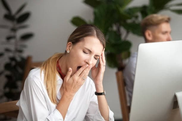 Woman at desk in office experiencing fatigue or stress, hand to face, with colleague working at computer in background among plants