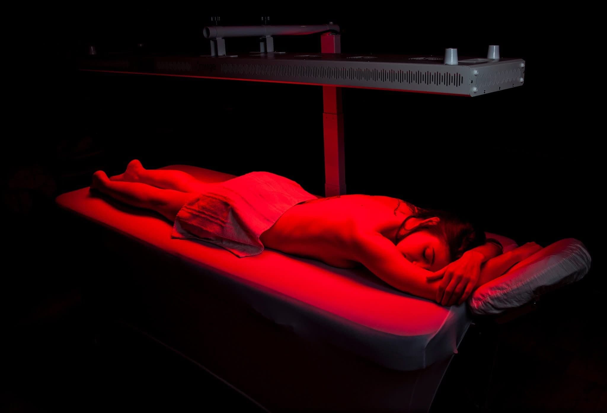 Red-lit sculptural installation of a reclining figure on a platform beneath an overhead heat lamp, suggesting therapeutic or wellness treatment